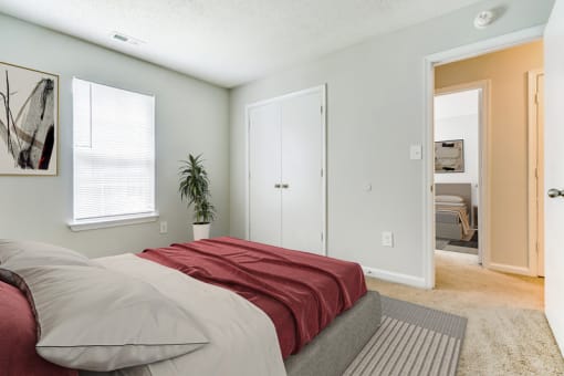 A virtually staged bedroom with white walls, carpet throughout, double doors opening to a closet, a single window with blinds, and an entry door leading to the hallway. It is staged with a gray area rug, a queen bed with burgundy and gray bedding, a green plant in the corner of the room and framed artwork hanging on one wall. The 2nd bedroom across the hall is in  the background.