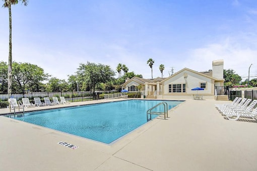 Swimming pool and sundeck with tables, chairs, and loungers. The perimeter fence is lined with trees and the exterior of the clubhouse.