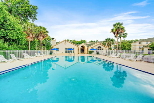 Swimming pool and sundeck with tables, chairs, and loungers. The perimeter fence is lined with trees and the exterior of the clubhouse.