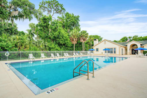 Swimming pool and sundeck with tables, chairs, and loungers. The perimeter fence is lined with trees and the exterior of the clubhouse.