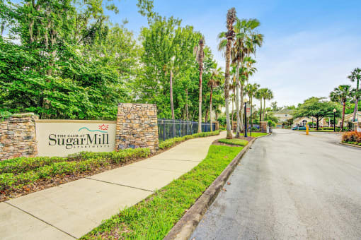Stone pillars on either side of a beige insert that reads The Club at Sugar Mill Apartments. The sign is on the main street before the access gates to the community, in front of mature trees and manicured landscaping.