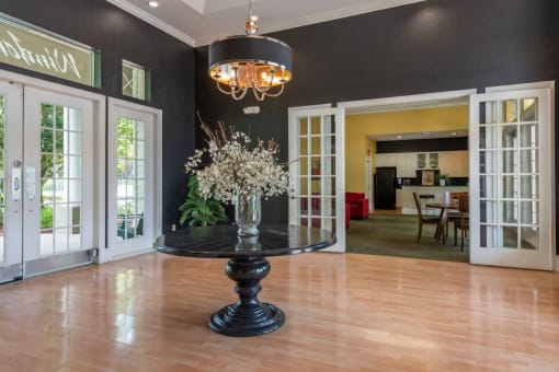 Clubhouse Foyer with a Table in the Middle with A Vase and Flowers on it Hardwood Floors Large Windows and Open Doors Leading to Clubhouse Seating Area with Kitchen