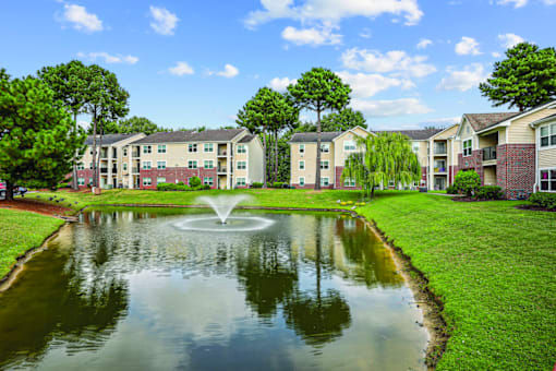 A pond with a fountain in the middle of a grassy area in front of apartment buildings.