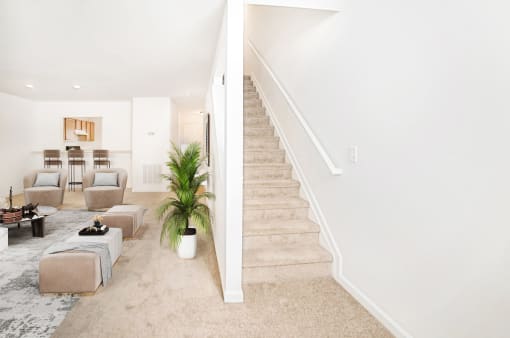 Virtually staged living room with carpet, grey accent chairs, and glass coffee table. Kitchen and stairs in background