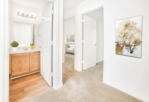 Carpeted bedroom looking towards bathroom with wood flooring and vanity. Virtually staged bathroom with décor and gold painting