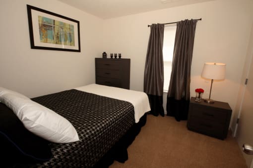 Bedroom with carpet flooring, dresser, night stand, desk lamp, wall art, and large window with curtains