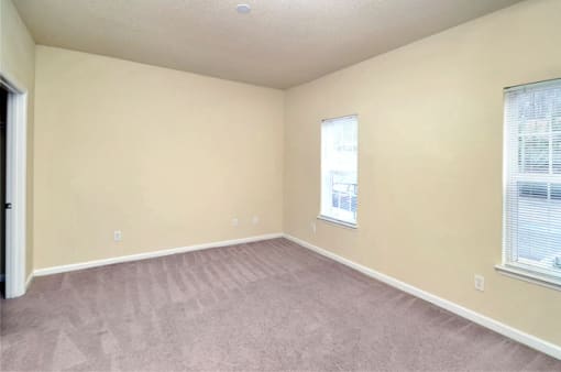 A vacant bedroom with tan carpet and beige walls. Two windows rest along the right side wall.