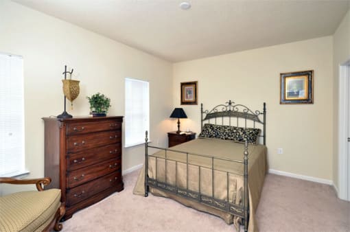 Bedroom with tan carpet, beige walls, and ceilings. The bed has a night stand to the left of the bed with a lamp and window.