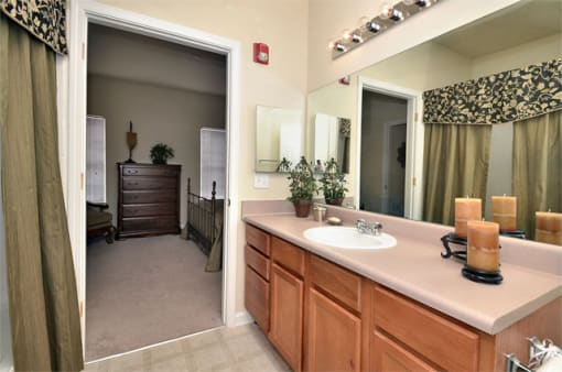 Bathroom with brown cabinets, sink, shower, and toilet. A large mirror is on the back wall with Hollywood lighting above.