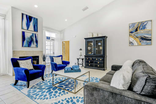 Clubhouse furniture with gray couch and white pillows, blue fabric chairs with pillows, a cabinet in the background with decor and abstract art on walls