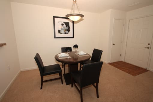 The furnished dining room has hardwood style flooring by the entry door, and carpet throughout the rest. There is a round wooden table with four black chairs positioned around it. There is a place setting for two on top of the table.