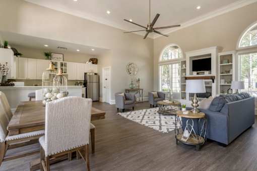 Clubhouse lounge with hardwood style flooring, couch, chairs, coffee table, rug, picnic table, fire place, book shelves, television, multi speed ceiling fan, and kitchen in the background
