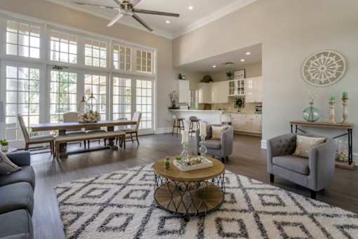 Clubhouse lounge with couch, chairs, coffee table, rug, picnic table, multi speed ceiling fan, large windows for natural lighting, and clubhouse kitchen in the background