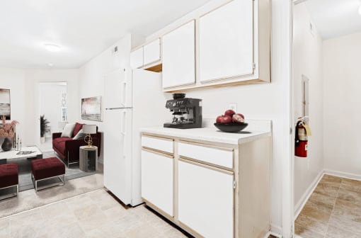 Virtually staged kitchen with tile floors, white appliances, and wood cabinets. Living room in background with maroon accent furniture.