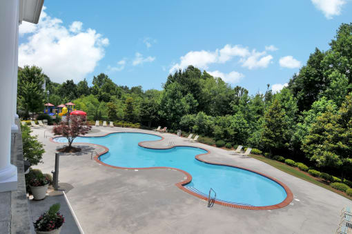 Community pool with pool furniture throughout. The clubhouse is in the foreground, trees surround the pool deck.