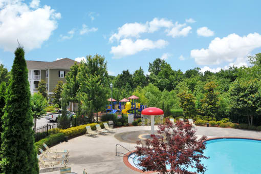 Pool deck with pool furniture throughout. Trees surround the pool deck with a splash pad and play area in the background.