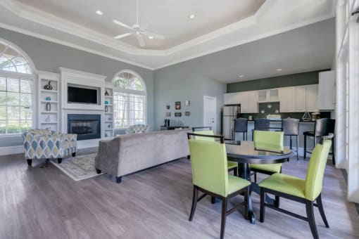 Clubhouse Seating Area with Large Windows on either side of Fireplace with TV and Shelves Surrounding Fireplace Multi Colored Accent Chairs Grey Couch Dining Table and Green Chairs and Clubhouse Kitchen with White Cabinets and Stainless Steel Appliances