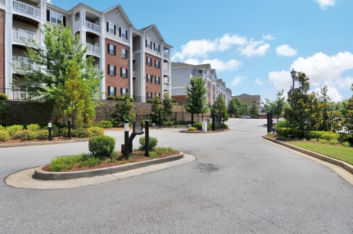 Community gated entrance with black fence surrounding entryway. Apartment buildings lie on the left side with mature trees.