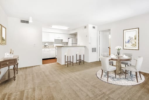 Vacant staged living room with wall-to-wall carpet. Kitchen in the background with white cabinets and appliances. Dining table in living room with buffet table by the wall and barstools by the breakfast bar.