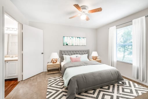 A staged bedroom with carpet, a ceiling fan, window, and an en suite bathroom. Full bed with tufted headboard on top of a rug and two night stands, and art above the bed.