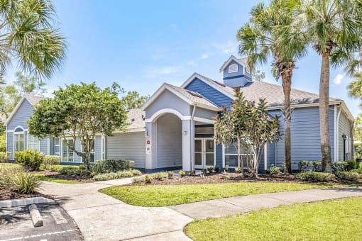 The exterior of a leasing office with trees, palm trees, a sidewalk, and grass areas.
