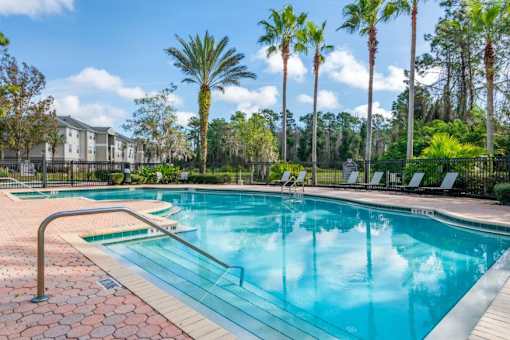 Fenced in Swimming Pool and Sun Deck with Lounge Chairs with Treeline and Building Exteriors in the Background