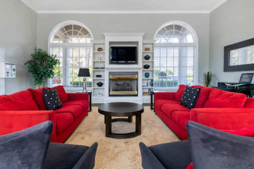 Clubhouse Seating Area with Large Windows Red Couches Wood Coffee Table Grey Arm Chairs with Fireplace and Decorative Plant Next to Mirror Mounted on the Wall Above a Table