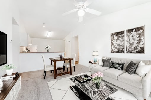 Staged living room with a dining room with carpeted flooring, a ceiling fan, mounted tv, with view of kitchen in the background.
