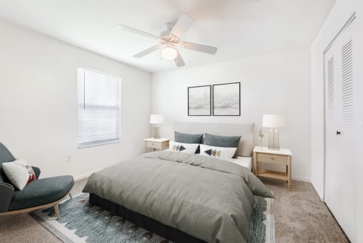 A staged bedroom with carpet, and a ceiling fan, window. Full bed with headboard on top of a rug and two nightstands, and art above the bed.