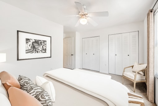 A staged bedroom with carpet, and a ceiling fan, window. Full bed with tufted headboard on top of a rug and art by the bed.