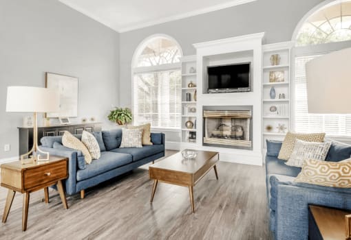 Clubhouse interior with blue couches, brown accent tables, fireplace, wall mounted television, gray walls with large windows