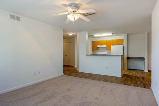 Carpeted living room with white walls and ceiling fan with the kitchen and built in desk in the background