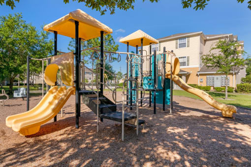 Playground jungle gym and playground slides in a bed of mulch with trees, and building exteriors in the background