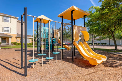 Playground jungle gym and playground slides in a bed of mulch with trees, and building exteriors in the background