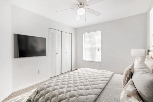 A staged bedroom with carpet, and a ceiling fan, window. Full bed with headboard and art above the bed with a mounted tv.