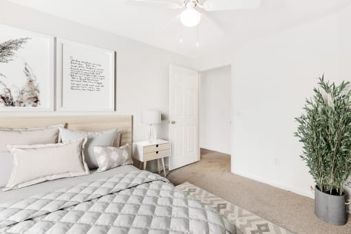 A staged bedroom with carpet, and a ceiling fan, window. Full bed with headboard on top of a rug and a nightstand, and art above the bed.