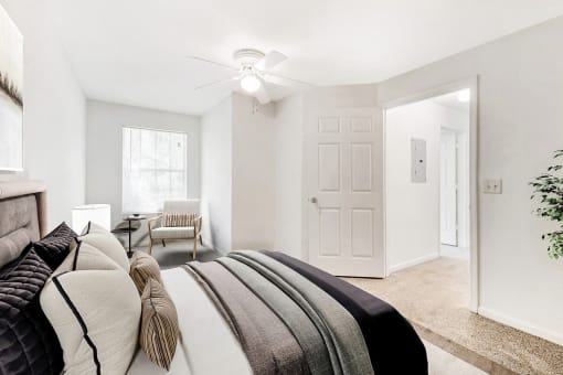 A staged bedroom with carpet, and a ceiling fan, window. Full bed with headboard on top of a rug and a nightstand, and art above the bed.