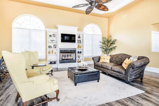 Clubhouse interior with fire place, wall mounted television, wood plank flooring, large windows, couch and chairs with ottoman and ceiling fan