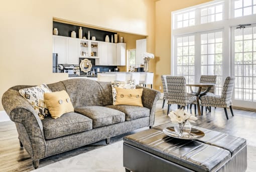 Clubhouse interior with gray couch, ottoman, accent rug and clubhouse kitchen in background with bar top and dining table with chairs