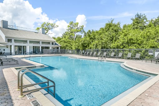 Swimming pool with large tanning deck, table with umbrella, lounge chairs, clubhouse in background surrounded by native landscaping