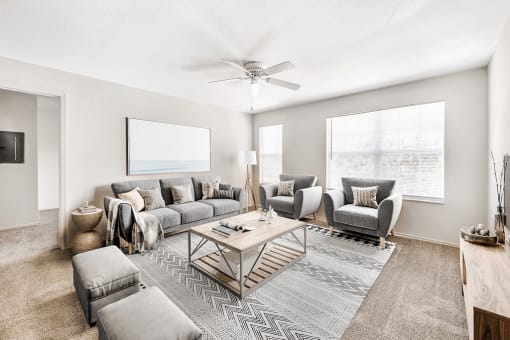 Virtually staged living room with carpet, accent rug, gray couches and seating, tan coffee table, ceiling fan, large windows