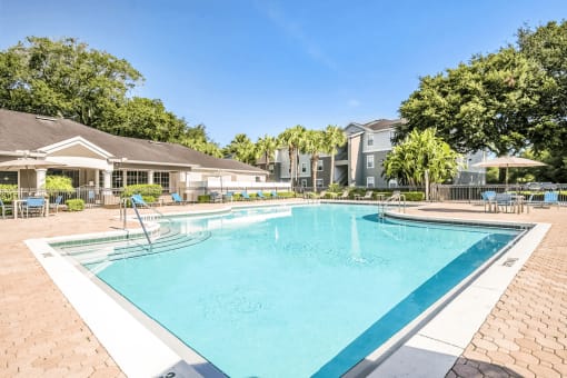 Swimming pool with large tanning deck, blue chairs, table with umbrella, clubhouse in background and surround native landscape
