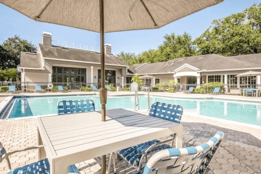 Swimming pool with large tanning deck, blue chairs, table with umbrella, clubhouse in background and surround native landscape