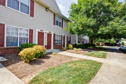 The apartment building exteriors are cream to tan in color with white trim and red shutters and doors. There are sidewalks leading to individual apartment doors, grass in areas between. the sidewalks, and large mature tree outside.