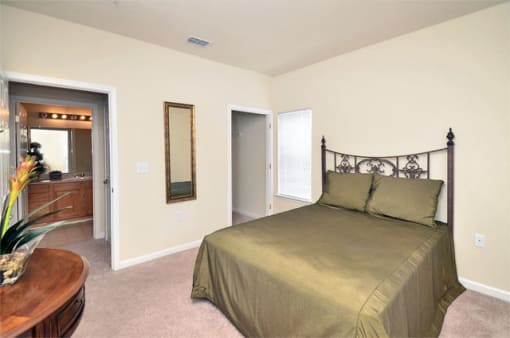 Bedroom with tan carpet, beige walls, and ceilings. The bed has a night stand to the left of the bed with a lamp and window.