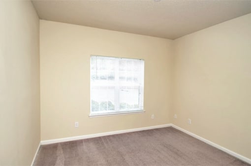 A vacant bedroom with tan carpet, beige walls, and ceilings. A single window is centered on the back wall.