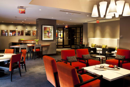 Clubroom seating area. Multiple orange accent chairs with white coffee table. Kitchen in background with bar seating and additional seating behind bar with black accent chairs.