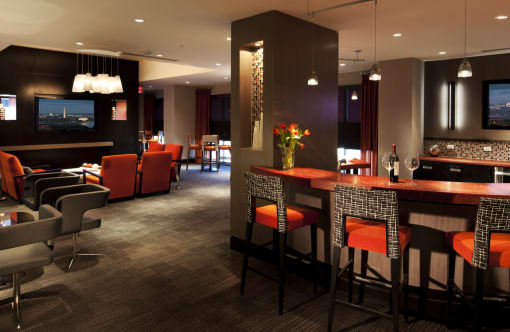 Clubroom kitchen. Bar area with four bar stools overlooking kitchen with tv on wall. Orange and wood accent colors with artwork on wall. Additional seating in background, 4 top table and orange accent chairs next to tv
