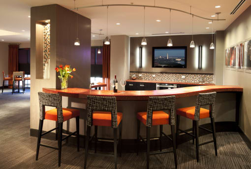 Clubroom kitchen. Bar area with four bar stools overlooking kitchen with tv on wall. Orange and wood accent colors with artwork on wall. Additional seating in background, 4 top table.