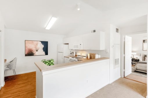 Virtually staged kitchen with white appliances, wood flooring, gas stove, white cabinetry, computer chair, wall art